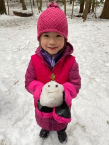 A smiling girl holds a small snowball with a face made of ferns and sticks