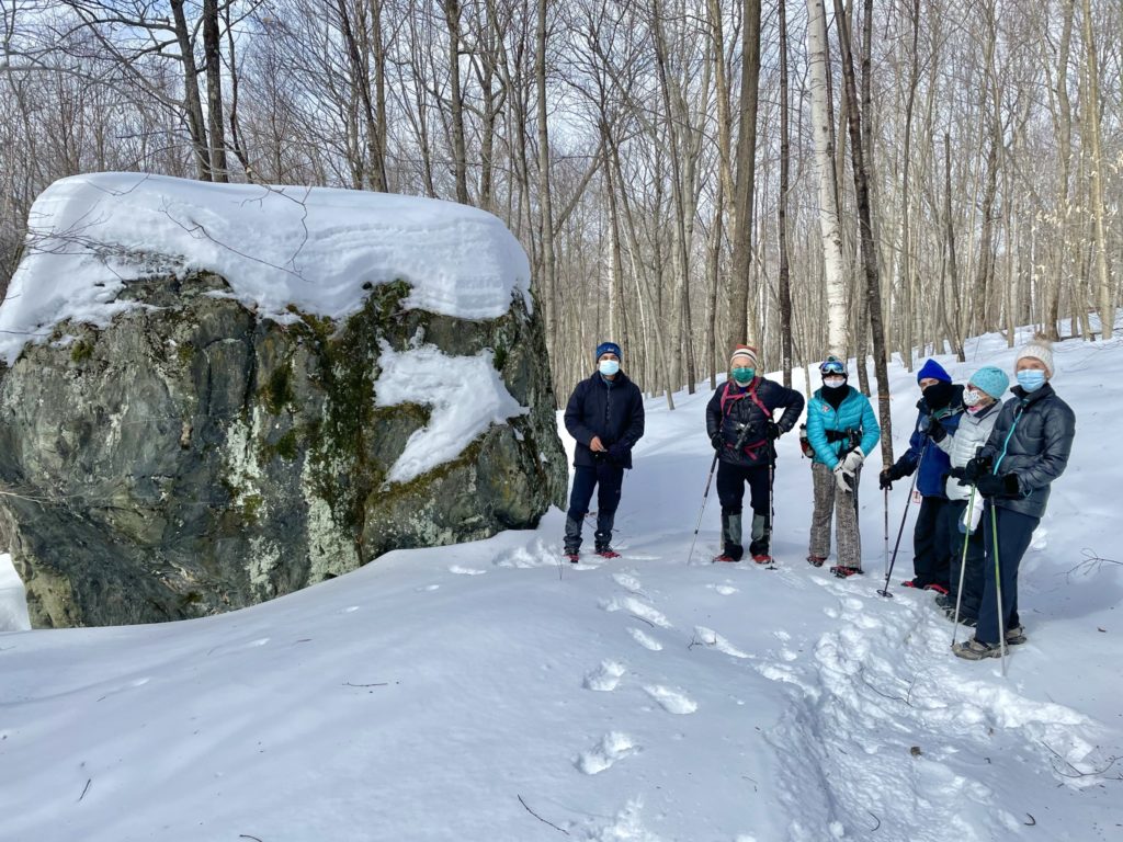 A group of hikers stands next to the big boulder at the Britton Forest, wearing masks and winter clothing. 